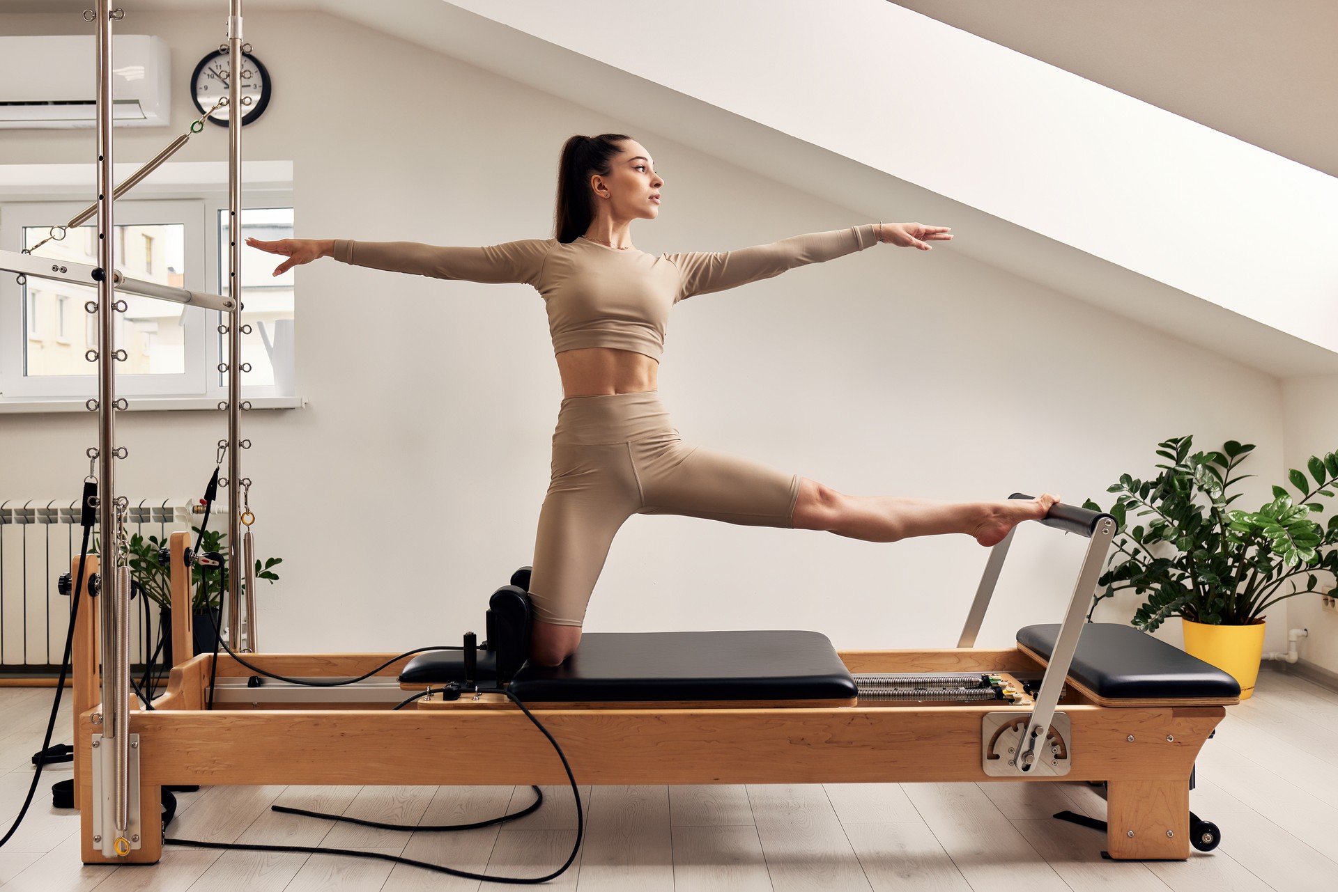 Young girl is doing Pilates on a reformer bed in a bright studio. Slender brunette in a beige bodysuit does exercise to train balance and stretch the muscles of her legs. Healthy lifestyle concept.